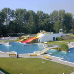Freibadkomplex mit mehreren Becken, Wasserrutschen, Springbrunnen, Rasenflächen und Bänken, umgeben von Bäumen unter klarem Himmel.