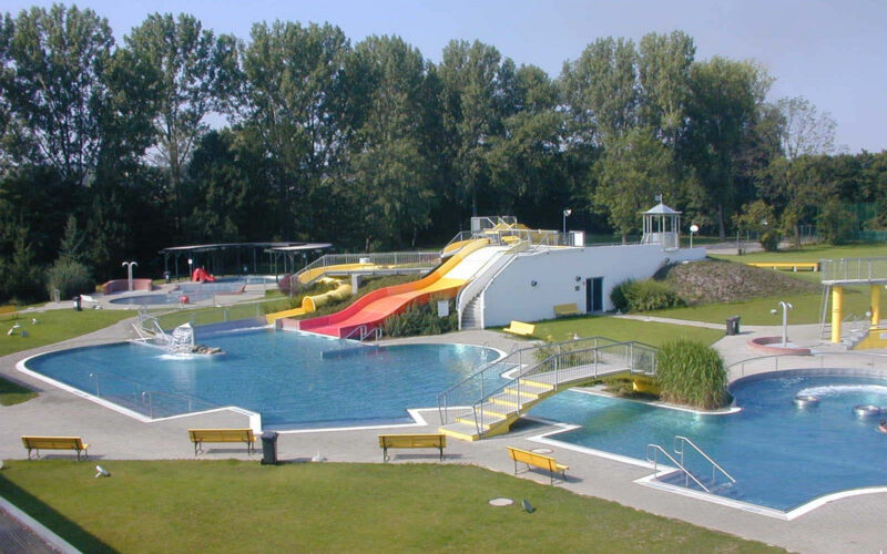 Freibadkomplex mit mehreren Becken, Wasserrutschen, Springbrunnen, Rasenflächen und Bänken, umgeben von Bäumen unter klarem Himmel.