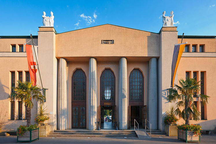 Symmetrisches Gebäude mit hohen Bogenfenstern, großen Säulen, Palmen, Fahnen auf jeder Seite und weißen Statuen auf dem Dach unter einem klaren blauen Himmel.