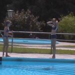Zwei Kinder in Badeanzügen stehen am Rande eines Schwimmbeckens, wobei eines von ihnen seine Schwimmbrille zurechtrückt. Im Hintergrund sind ein rundes Planschbecken und Grünflächen zu sehen.