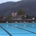 Freibad mit Bahnmarkierungen im Vordergrund, mit Gebäuden, Bäumen und Hügeln im Hintergrund unter einem bewölkten Himmel.