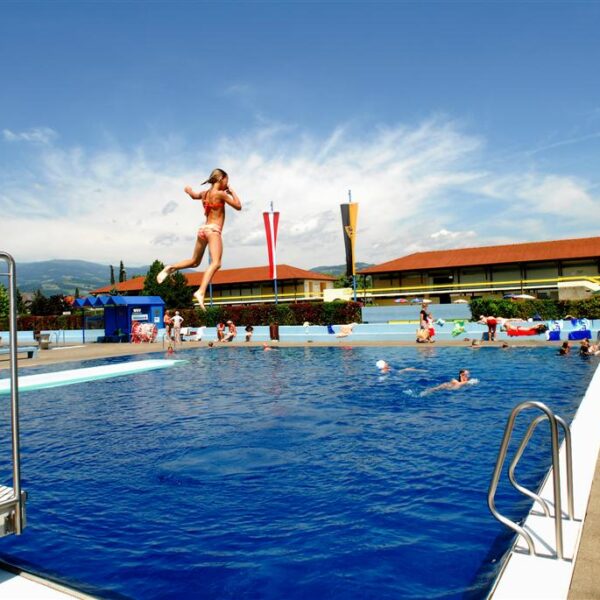 Eine Person springt in ein Freibad, während andere an einem sonnigen Tag um den Pool herum schwimmen und sich entspannen.