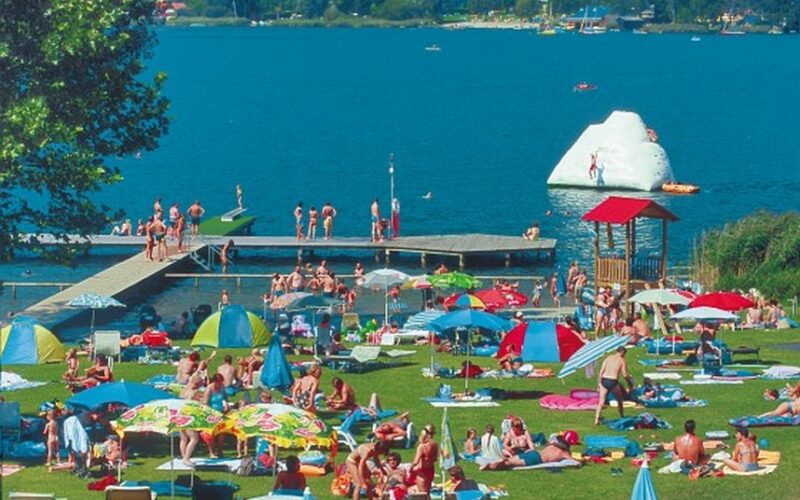 Belebter Strand am See mit Menschen, die sich im Gras sonnen, bunten Sonnenschirmen, einem Steg und Schwimmern; ein aufblasbarer Eisberg schwimmt im Wasser.