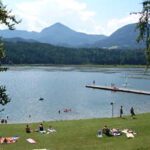 Menschen sonnen sich und schwimmen an einem Seeufer mit einem grasbewachsenen Ufer, einem hölzernen Steg und Bergen im Hintergrund unter einem teilweise bewölkten Himmel.