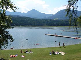 Menschen sonnen sich und schwimmen an einem Seeufer mit einem grasbewachsenen Ufer, einem hölzernen Steg und Bergen im Hintergrund unter einem teilweise bewölkten Himmel.
