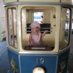 Ein kleines Kind mit einer rosa Mütze sitzt in einer blau-gelben Straßenbahn mit der Aufschrift "Festplatz" und der Nummer 19 und schaut zur Seite.
