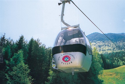 Eine weiße Seilbahn mit "Petzen"-Logo fährt über eine grüne, bewaldete Landschaft mit Bergen im Hintergrund.