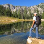 Ein Mann mit Rucksack steht auf einem Felsen an einem klaren See, umgeben von Bäumen und Bergen unter einem blauen Himmel.