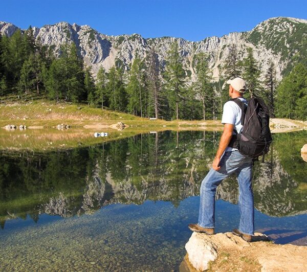 Ein Mann mit Rucksack steht auf einem Felsen an einem klaren See, umgeben von Bäumen und Bergen unter einem blauen Himmel.