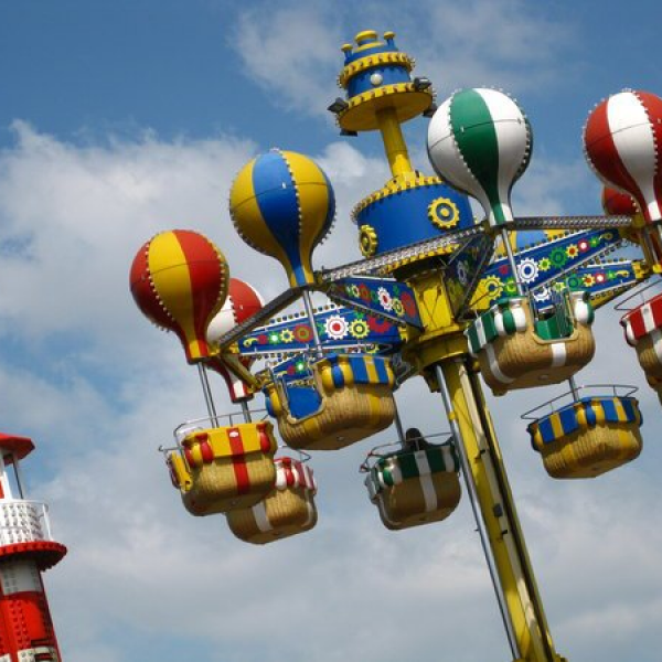 Buntes Fahrgeschäft im Vergnügungspark mit Sitzen in Form eines Heißluftballons, die sich in der Nähe eines rot-weiß gestreiften Turms vor einem teilweise bewölkten Himmel drehen.