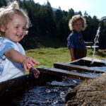 Ein kleines Kind lächelt, während es mit Wasser aus einem Wasserhahn im Freien spielt, während ein anderes Kind im Hintergrund auf einem grasbewachsenen Hügel steht.
