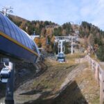 Ein Skilift befördert die Gondeln einen grasbewachsenen, baumbestandenen Hang hinauf, an dem ein Holzzaun entlangführt.
