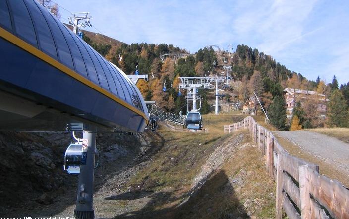 Ein Skilift befördert die Gondeln einen grasbewachsenen, baumbestandenen Hang hinauf, an dem ein Holzzaun entlangführt.