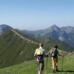Zwei Wanderer mit Rucksäcken und Wanderstöcken gehen entlang eines grasbewachsenen Bergrückens mit Gipfeln und einem Gebäude im Hintergrund unter einem klaren Himmel.
