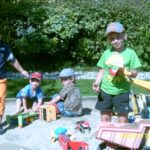 Vier kleine Kinder spielen mit Spielzeuglastwagen und Baufahrzeugen in einem Sandkasten im Freien, umgeben von Grün und Sonnenlicht.
