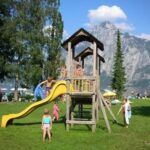 Kinder spielen auf einem hölzernen Spielplatz mit einer gelben Rutsche in einem grasbewachsenen Park, während sich die Menschen entspannen und die Berge im Hintergrund zu sehen sind.
