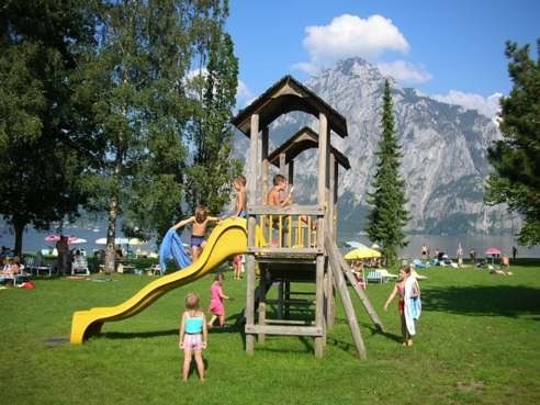 Kinder spielen auf einem hölzernen Spielplatz mit einer gelben Rutsche in einem grasbewachsenen Park, während sich die Menschen entspannen und die Berge im Hintergrund zu sehen sind.