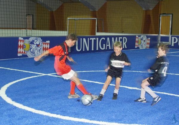 Drei Jungen spielen Fußball auf einem blauen Hallenfeld; einer in einem roten Trikot schießt den Ball, während zwei andere in schwarzen Trikots versuchen, ihn zu verteidigen.
