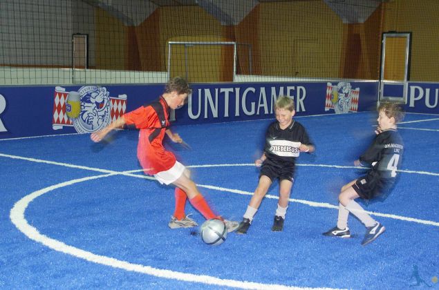 Drei Jungen spielen Fußball auf einem blauen Hallenfeld; einer in einem roten Trikot schießt den Ball, während zwei andere in schwarzen Trikots versuchen, ihn zu verteidigen.