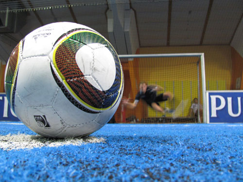 Nahaufnahme eines Fußballs auf einem blauen Hallenfeld, mit einem unscharfen Torwart, der auf das Tor im Hintergrund zuläuft.