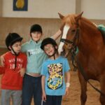 Drei Kinder mit Helmen stehen neben einem braunen Pferd in einer Reithalle und lächeln in die Kamera.