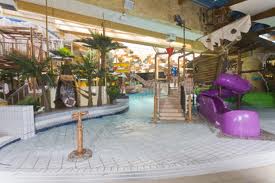 Indoor-Wasserpark mit einem flachen Becken, Palmen, Spielgeräten, einer Seilbrücke und einer lila Rutsche.