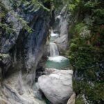 Ein kleiner Wasserfall fließt zwischen felsigen Klippen in einen klaren, türkisfarbenen Pool, der von üppiger grüner Vegetation umgeben ist.
