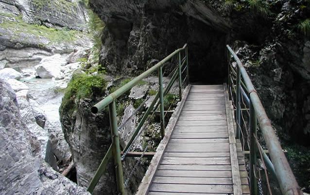 Eine schmale Holzbrücke mit Metallgeländer führt über eine felsige Schlucht, durch die ein Bach fließt.