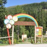 Eingang zum Erlebnispark Katschhausen mit einem Holzschild mit einem Regenbogen und Cartoon-Tieren, umgeben von Bäumen und einer Waldlandschaft.