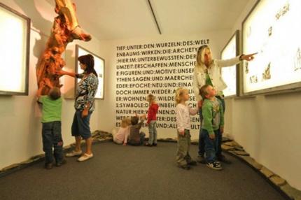 In einem hell erleuchteten Museumsraum mit Text an der Rückwand betrachten Kinder und Erwachsene Schautafeln und eine Baumausstellung.