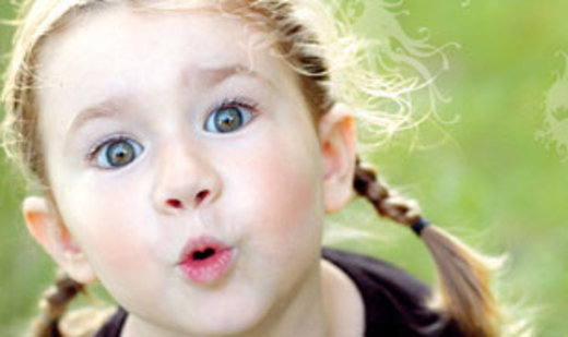 Kind mit hellbraunem Haar in Zöpfen und großen grünen Augen, die mit dem Mund ein "O" formen, im Freien vor einem verschwommenen grünen Hintergrund.