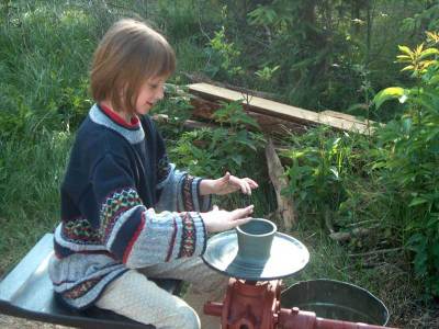 Ein Kind formt Ton auf einer Töpferscheibe im Freien, umgeben von Grünpflanzen und Holzstämmen.