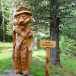 Holzstatue eines bärtigen Mannes mit Krone und Stab in der Hand, der in einem Wald neben einem Schild mit der Aufschrift "König Waldgeist" steht.