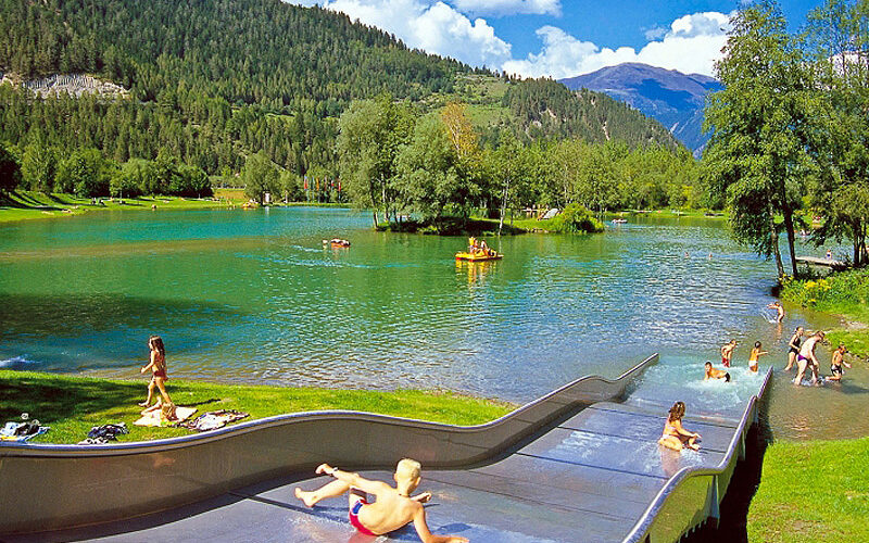 Kinder benutzen eine Wasserrutsche, die in einen klaren See führt, der von Bäumen und Bergen unter einem blauen Himmel mit Wolken umgeben ist.