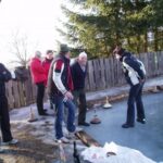 Eine Gruppe von Menschen steht an einem Holzzaun und spielt ein Spiel auf einer vereisten Fläche im Freien, mit Bäumen im Hintergrund. Einige Curlingsteine und Flaschen liegen auf dem Boden.