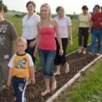 Eine Gruppe von Menschen, darunter Kinder und Erwachsene, läuft barfuß über einen schmalen Weg im Freien, der mit kleinen Steinen oder Mulch bedeckt ist.