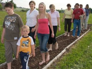 Eine Gruppe von Menschen, darunter Kinder und Erwachsene, läuft barfuß über einen schmalen Weg im Freien, der mit kleinen Steinen oder Mulch bedeckt ist.