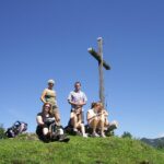 Fünf Menschen sitzen und stehen auf einer grasbewachsenen Anhöhe neben einem großen Holzkreuz unter einem strahlend blauen Himmel.