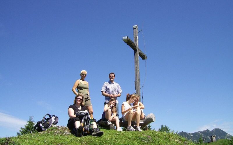 Fünf Menschen sitzen und stehen auf einer grasbewachsenen Anhöhe neben einem großen Holzkreuz unter einem strahlend blauen Himmel.