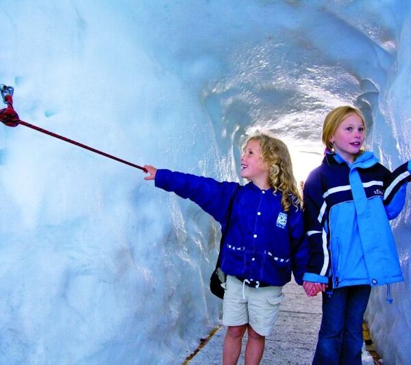 Zwei Kinder in blauen Jacken gehen durch einen Eistunnel, halten sich an den Händen und berühren ein rotes Seil an der eisigen Wand.