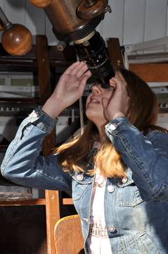 Eine Person in einer Jeansjacke schaut im Innenbereich durch das Okular eines großen Teleskops.