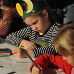 Ein Kind, das eine Papierkrone mit Mond und Sternen trägt, malt mit Buntstiften an einem Tisch zusammen mit anderen Kindern.