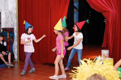 Drei Kinder mit bunten Zipfelmützen und roten Clownsnasen treten auf der Bühne vor einem roten Vorhang auf, während andere Kinder sitzen und zusehen.