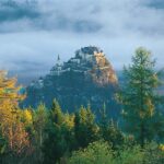 Ein Schloss thront auf einem felsigen Hügel, umgeben von Bäumen und Nebel, und im Hintergrund sind schwach die Berge zu erkennen.