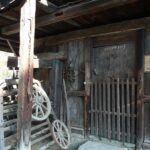 Holzschuppen mit rustikalen Werkzeugen und Geräten, einer vergitterten Holztür und einem Schild mit der Aufschrift "2. Samalmador 19 St. Johann/Verns". Das Sonnenlicht wirft Schatten auf das verwitterte Gebäude.