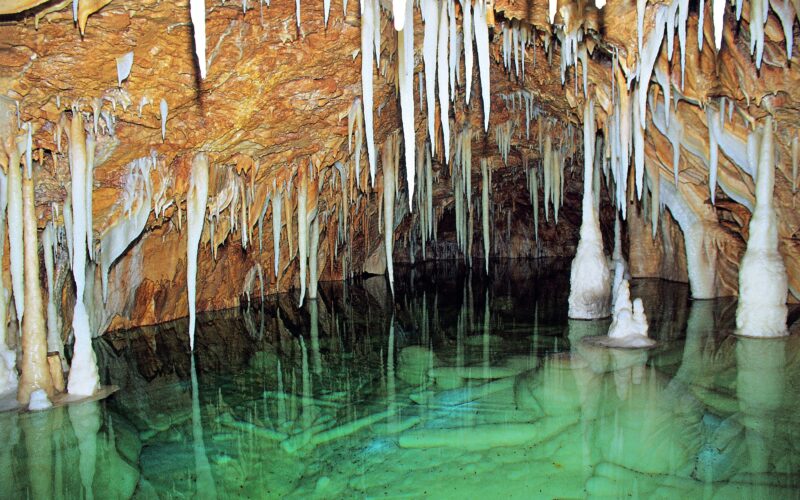 Unterirdische Höhle mit hängenden Stalaktiten, stehenden Stalagmiten und klarem grünen Wasser, in dem sich die Formationen spiegeln.