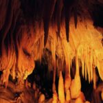Ein Höhleninneres mit zahlreichen orangefarbenen Stalaktiten, die von der Decke hängen, und Stalagmiten, die aus dem Boden ragen, beleuchtet von warmem Licht.