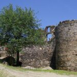 Die steinernen Ruinen eines alten Gebäudes mit einem zylindrischen Turm und einem Baum, der in der Nähe wächst, stehen unter einem klaren blauen Himmel.