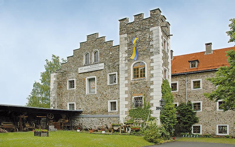 Steingebäude mit dekorativen weißen Akzenten, historischen Schildern, einem blau-gelben Emblem und einem Außenbereich mit alten Werkzeugen und Geräten auf der linken Seite.