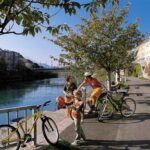 Drei Personen mit Fahrrädern und ein Kind stehen auf einem von Bäumen gesäumten Uferweg, im Hintergrund sind Gebäude und eine Brücke zu sehen.